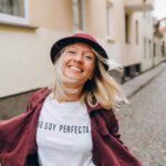 Blonde woman smiling widely in a hat and coat on a cobblestone street, embodying joy and freedom.